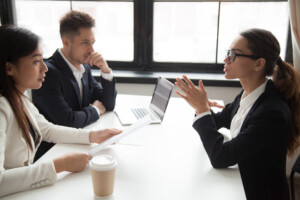 Three people in business attire sit at a table during an interview. Two are on one side with a laptop and paper, while the third, gesturing with hands, sits opposite them. 