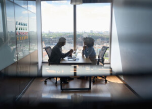 Two people sit across from each other at a table in a modern office with a large window. The view outside shows a cityscape under a cloudy sky. 