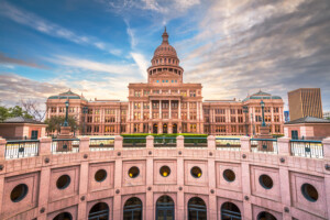 The Texas State Capitol building in Austin, with a large dome and ornate facade, stands under a dramatic, partly cloudy sky, viewed from the sunken rotunda in the foreground.