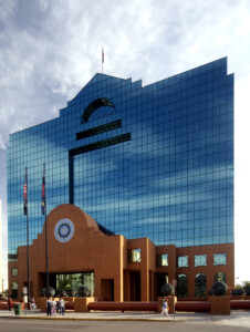 El Paso courthouse with glass facade reflecting the sky, featuring a central section with red brick, a round decorative emblem, and two flagpoles in front. People walk on the sidewalk nearby.