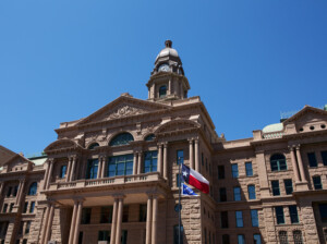 Fort Worth courthouse with columns and a central dome under a clear blue sky, featuring the Texas state flag and another flag flying in front.