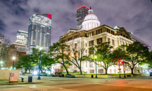 Historic Houston Texas courthouse with white dome is illuminated at night, surrounded by trees and modern skyscrapers under a cloudy sky in a cityscape. Cars are parked along the street in the foreground.