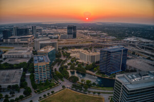 Aerial view of Plano Texas cityscape at sunset, with tall office buildings, highways, green spaces, and a small pond, as the sun sets on the horizon and casts an orange glow over the scene.