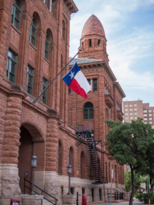 A Texas flag waves in front of a San Antonio courthouse with arched windows and a tower. Trees and another tall building are in the background on a partly cloudy day.