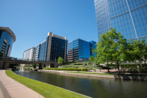 A canal runs alongside a sidewalk with modern glass office buildings and green trees under a clear blue sky on a sunny day in The Woodlands, Texas