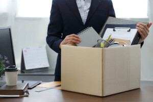 A lawyer packs office supplies, notebooks, and a clipboard into a cardboard box, suggesting resignation or job loss. A desk, laptop, and calendar are visible in the background.