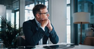 Attorney in a suit and glasses sits at a desk with his hands under his chin, looking thoughtfully out the window in a modern office setting. 