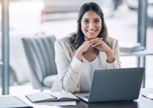 A female lawyer in a light-colored blazer smiles at the camera while sitting at a desk with a laptop