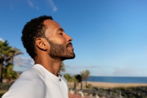 A man with closed eyes stands outdoors, facing the sun, appearing calm and relaxed.