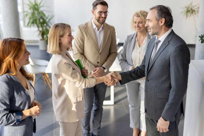 Five legal professionals in formal attire are standing in an office. Two of them, a man and a woman, are shaking hands and smiling, while the others watch and smile in the background. 
