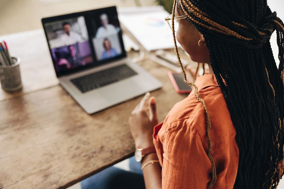 A woman with braided hair sits at a wooden desk, participating in a video call on her laptop with four people visible on the screen. Pens, a phone, and papers are on the desk.